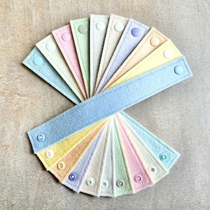 May include: A collection of pastel-colored felt bands, each with a snap closure. The bands are arranged in a fan shape, showcasing a variety of soft hues including blue, pink, yellow, and green. The felt has a stitched border, and the snaps are in matching colors.