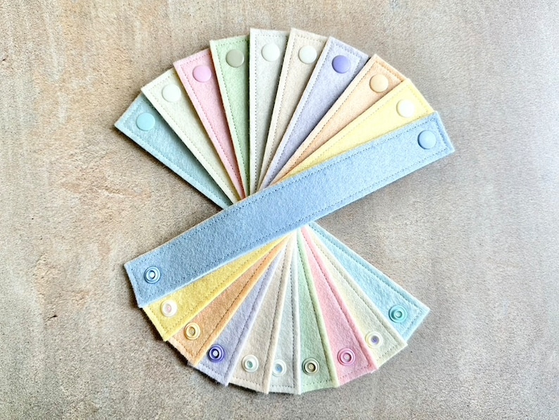May include: A collection of pastel-colored felt bands, each with a snap closure. The bands are arranged in a fan shape, showcasing a variety of soft hues including blue, pink, yellow, and green. The felt has a stitched border, and the snaps are in matching colors.