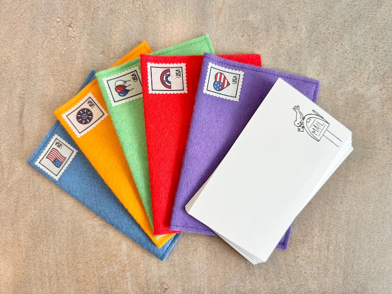 Peut inclure: Un ensemble de cinq enveloppes en feutrine bleue, jaune, verte, rouge et violette, chacune orn&eacute;e d'un timbre d&eacute;coratif avec le texte USA. Une pile de cartes rectangulaires blanches avec un dessin au trait noir d'un oiseau et d'une bo&icirc;te aux lettres est pos&eacute;e dessus.