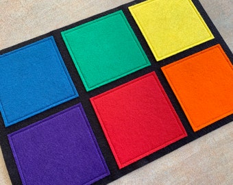Peut inclure: Un tableau de tri des couleurs en feutre, présentant six carrés vibrants en bleu, vert, jaune, orange, rouge et violet, chacun avec une bordure cousue. Les carrés sont posés sur un fond gris foncé.