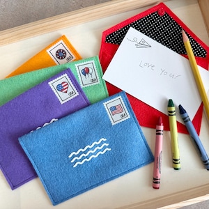 Peut inclure: Une collection d'enveloppes en feutrine color&eacute;e orange, verte, violette, bleue et rouge, avec des timbres d&eacute;coratifs portant l'inscription USA. Une note blanche avec le texte manuscrit Love you et un dessin d'avion en papier est accompagn&eacute;e de trois crayons et d'un crayon jaune.