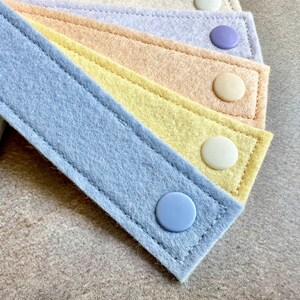 May include: Close-up of several pastel-colored felt straps with button closures. The straps are in shades of blue, yellow, peach, and lavender, with matching or contrasting buttons. The felt has a stitched border.