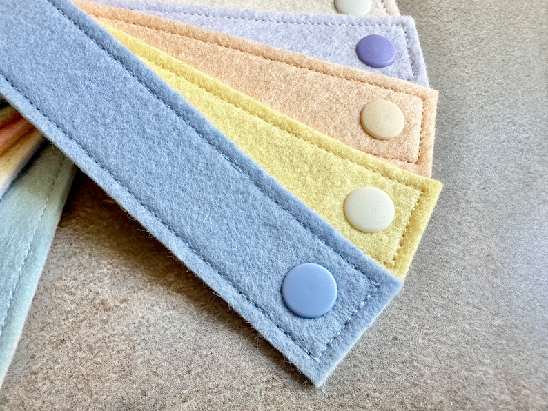 May include: Close-up of several pastel-colored felt straps with button closures. The straps are in shades of blue, yellow, peach, and lavender, with matching or contrasting buttons. The felt has a stitched border.