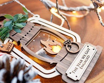 May include: Wooden Christmas ornament shaped like a lantern with a lit flame inside. The ornament has a small tag that says "The year we got engaged" and another tag that says "2022".