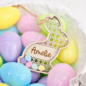 Peut inclure: Une découpe de lapin de Pâques en bois avec un motif tissé et le nom "Amelie" inscrit dessus. Le lapin est entouré d'œufs de Pâques en plastique de couleur pastel dans un panier blanc.