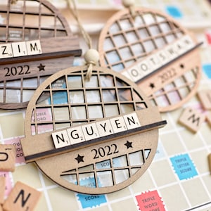 May include: Three wooden ornaments shaped like a circle with a grid pattern. Each ornament has a name spelled out with Scrabble tiles and the year 2022. The names are Azim, Nguyen, and Oots.