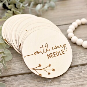 May include: A collection of light wooden circular discs, each engraved with the words "worth every NEEDLE" in a cursive font. A floral design is etched on the bottom left. White beads and greenery are in the background.
