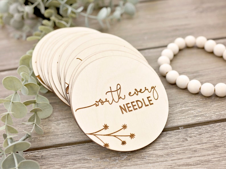 May include: A collection of light wooden circular discs, each engraved with the words "worth every NEEDLE" in a cursive font. A floral design is etched on the bottom left. White beads and greenery are in the background.