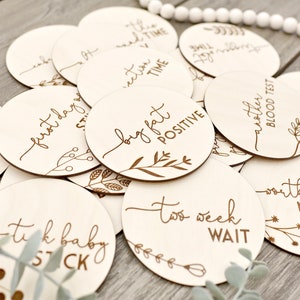 May include: A selection of circular wooden milestone markers, each engraved with phrases like "first day" and "big fat positive", alongside floral designs. Perfect for baby announcements and pregnancy milestones.