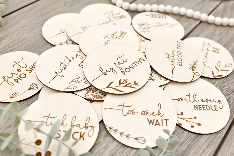 May include: A selection of circular wooden milestone markers, each engraved with phrases like "first day" and "big fat positive", alongside floral designs. Perfect for baby announcements and pregnancy milestones.