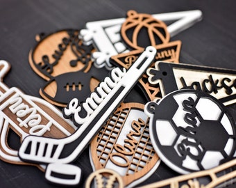Könnte beinhalten: Eine Sammlung personalisierter Holzornamente mit verschiedenen Sportmotiven, darunter ein Hockeyschläger, ein Golfschläger, ein Fußball, ein Basketball und ein Tennisschläger. Jedes Ornament ist mit einem Namen verziert, wie z. B. "Emma", "Jackson" und "Olivia".