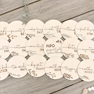 May include: A set of 18 wooden discs, each with laser-engraved text and botanical designs. The discs are inscribed with phrases such as "first pios," "official test day," and "big fat positive," for tracking milestones.