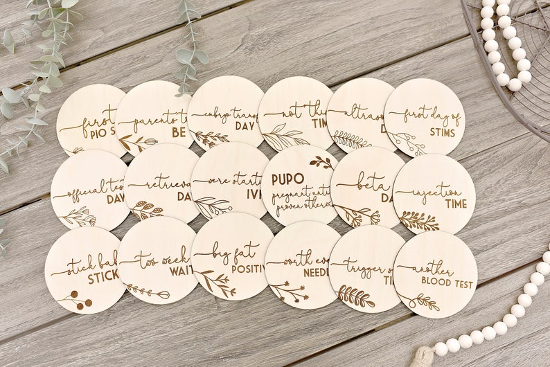 May include: A set of 18 wooden discs, each with laser-engraved text and botanical designs. The discs are inscribed with phrases such as "first pios," "official test day," and "big fat positive," for tracking milestones.