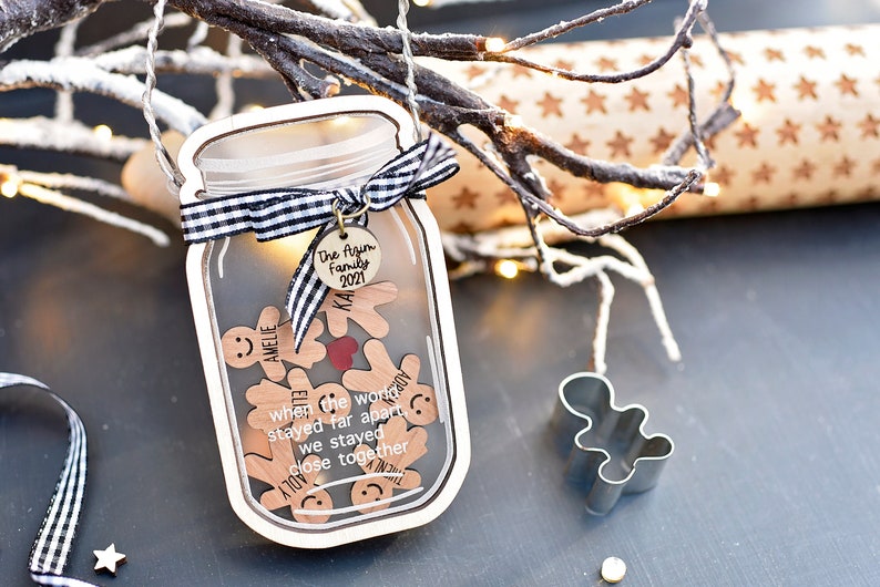 Puede incluir: Un adorno navide&ntilde;o de madera con forma de tarro de cristal con una cinta a cuadros en blanco y negro. El tarro contiene peque&ntilde;os hombres de jengibre de madera con nombres y el texto "when the world stayed far apart we stayed close together". El adorno tambi&eacute;n tiene una peque&ntilde;a etiqueta de madera con el texto "The ftzim Family 2021".