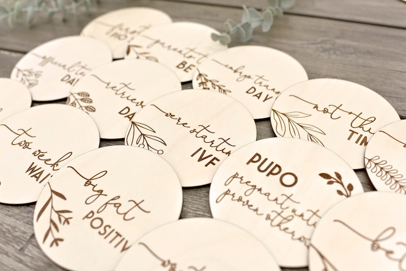 May include: A selection of light wooden discs, each engraved with text and botanical designs. Phrases such as "big fat positive" and "IVF" are visible. The discs are displayed on a wooden surface.