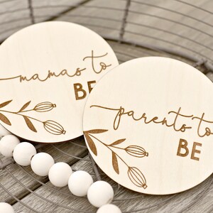 May include: Two circular wooden plaques with engraved text and floral designs. One says "mamas to BE," the other "parento to BE." The plaques are light wood and are displayed on a wire basket and white beads.