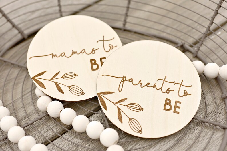 May include: Two circular wooden plaques with engraved text and floral designs. One says "mamas to BE," the other "parento to BE." The plaques are light wood and are displayed on a wire basket and white beads.