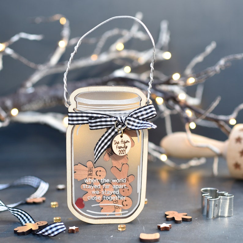 Puede incluir: Un adorno navide&ntilde;o de madera con forma de tarro de cristal con una cinta a cuadros en blanco y negro. El tarro est&aacute; lleno de hombres de jengibre y una etiqueta que dice "The Agim Family 2021". El adorno tiene el texto "When the world stayed far apart, we stayed close together" en la parte delantera.