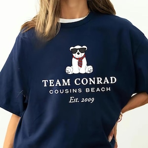 May include: Navy blue t-shirt with a white collar featuring a graphic of a polar bear wearing sunglasses and a scarf. The text "TEAM CONRAD, COUSINS BEACH, Est. 2009" is printed below the bear. The shirt has short sleeves.