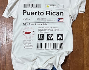 Pode incluir: Uma camiseta branca com um design gráfico preto e branco de um rótulo de envio. O rótulo diz "Puerto Rican" e inclui detalhes como "Local de origem" e "100% materiais orgânicos".