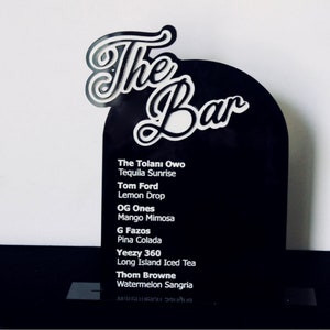 Puede incluir: Un letrero negro con texto blanco que dice "The Bar" en la parte superior. El letrero enumera una variedad de cócteles, incluyendo The Tolanı Owo, Tequila Sunrise, Tom Ford, Lemon Drop, OG Ones, Mango Mimosa, G Fazos, Pina Colada, Yeezy 360, Long Island Iced Tea, Thom Browne y Watermelon Sangria.