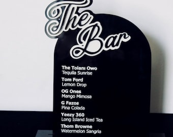 May include: Black sign with white text that reads "The Bar" at the top. The sign lists a variety of cocktails, including The Tolanı Owo, Tequila Sunrise, Tom Ford, Lemon Drop, OG Ones, Mango Mimosa, G Fazos, Pina Colada, Yeezy 360, Long Island Iced Tea, Thom Browne, and Watermelon Sangria.