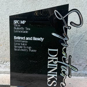 Puede incluir: Un letrero de acrílico negro con letras plateadas que enumeran las bebidas de autor: SFC MP Tilo's Butterfly Tea Lemonade, Retired and Ready Teremana Lime Juice Simple Syrup Strawberry Puree. El letrero dice "Signature Drinks" en una escritura estilizada.