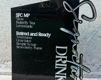 May include: Black acrylic sign with silver lettering listing signature drinks: SFC MP Tilo's Butterfly Tea Lemonade, Retired and Ready Teremana Lime Juice Simple Syrup Strawberry Puree. The sign reads "Signature Drinks" in a stylized script.