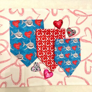 May include: Three dog bandanas with a shark and heart design. The bandanas are blue with white sharks and red hearts that say "Love Bites". One bandana is red with white "X" patterns.