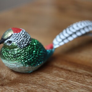 May include: A small, green and silver bird figurine with a red and white patterned head and a long, white and black feathered tail.