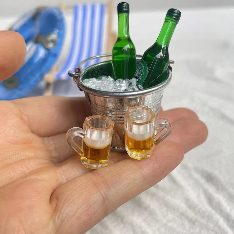 May include: Two miniature glass beer mugs filled with a light yellow liquid, next to a silver bucket filled with ice and two green glass bottles.