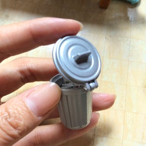 May include: A small, silver, miniature trash can with a hinged lid. The lid is open and the trash can has a ribbed texture.