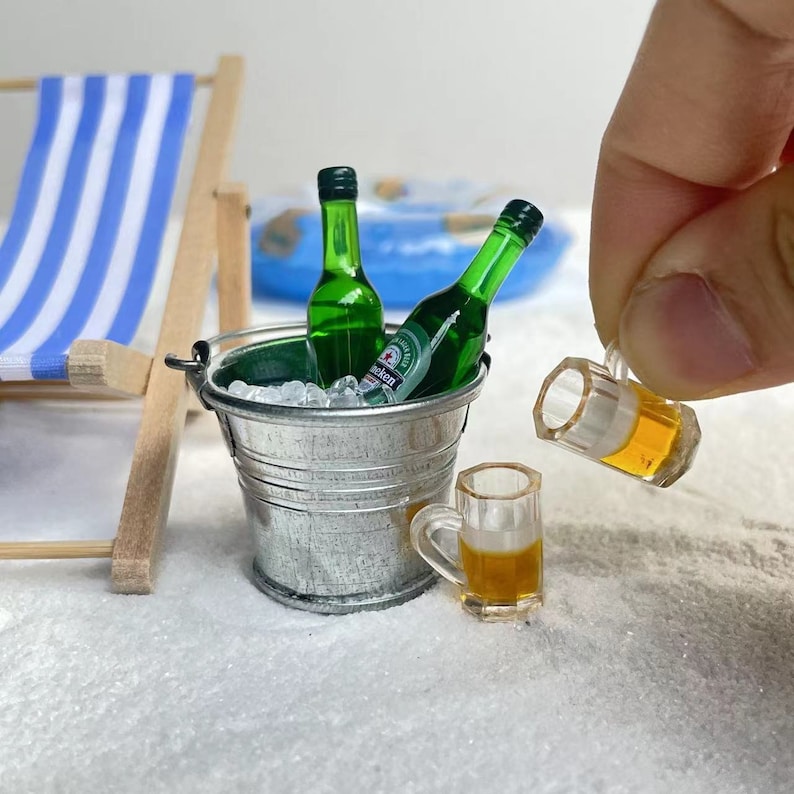 May include: A miniature metal bucket filled with ice and two green glass bottles. A miniature glass of beer is being poured from a miniature glass mug.
