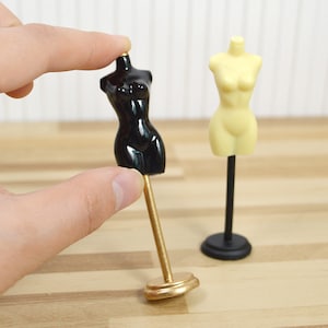 May include: Two miniature dress forms, one black and one cream, on stands. The black dress form has a gold stand, and the cream dress form has a black stand.