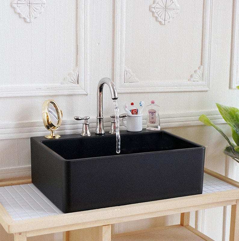 May include: A miniature black rectangular sink with a chrome faucet, a small gold mirror, and a cup with toothbrushes. The sink is on a wooden shelf with a white tile surface, set against a white wall.