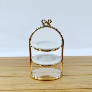 May include: A gold metal two-tiered serving stand with white ceramic plates. The stand has a bow detail at the top.