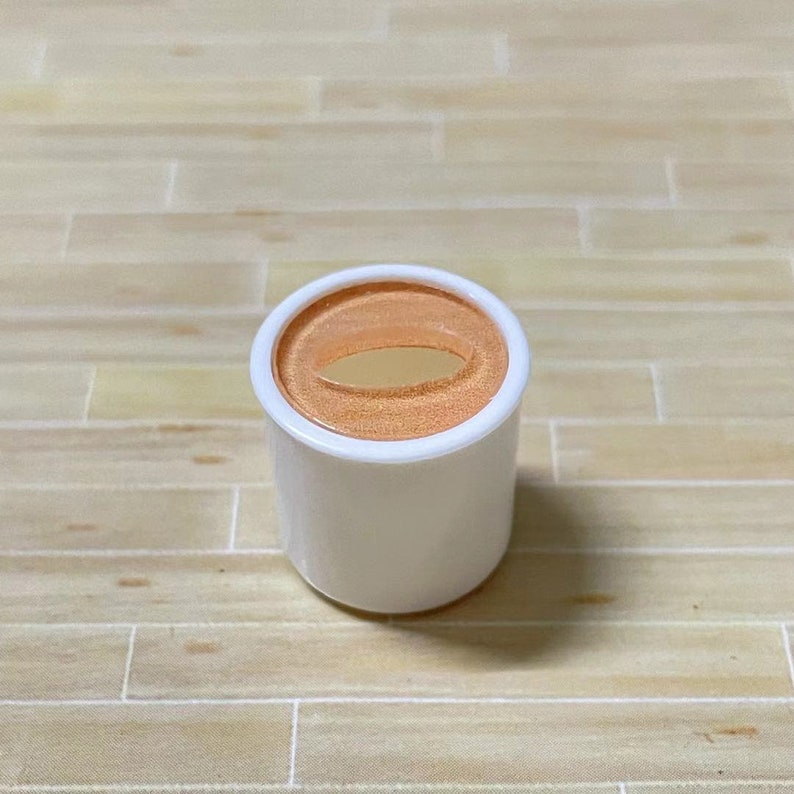 May include: A white plastic container with a round, orange lid. The lid has a small, oval-shaped opening in the center.