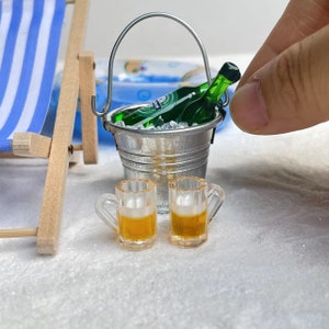 May include: Two miniature glass beer mugs with a green glass beer bottle in a silver bucket with ice. The bucket has a handle and is on a white surface.