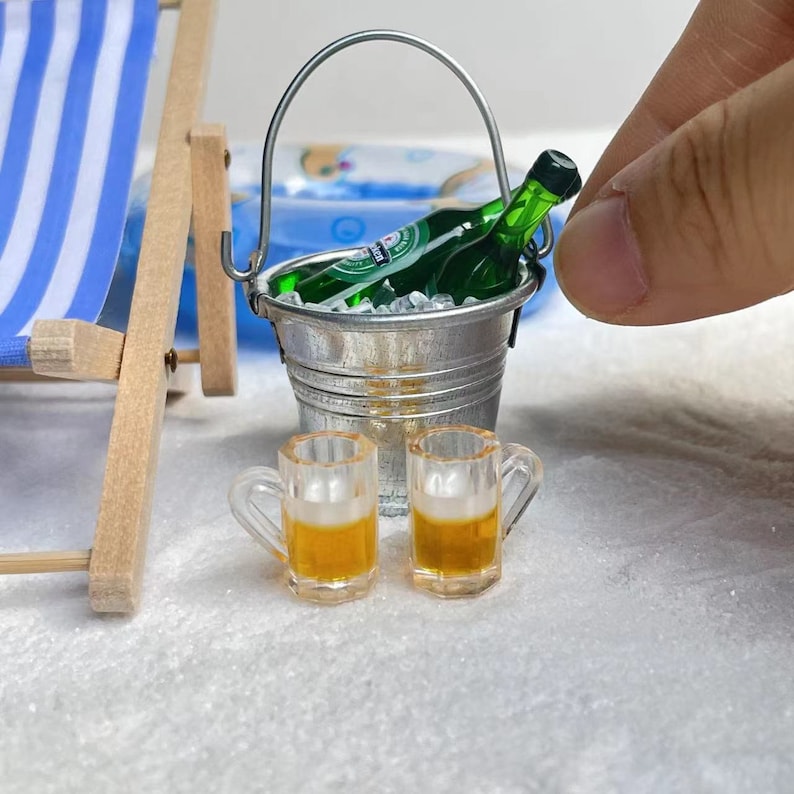 May include: Two miniature glass beer mugs with a green glass beer bottle in a silver bucket with ice. The bucket has a handle and is on a white surface.