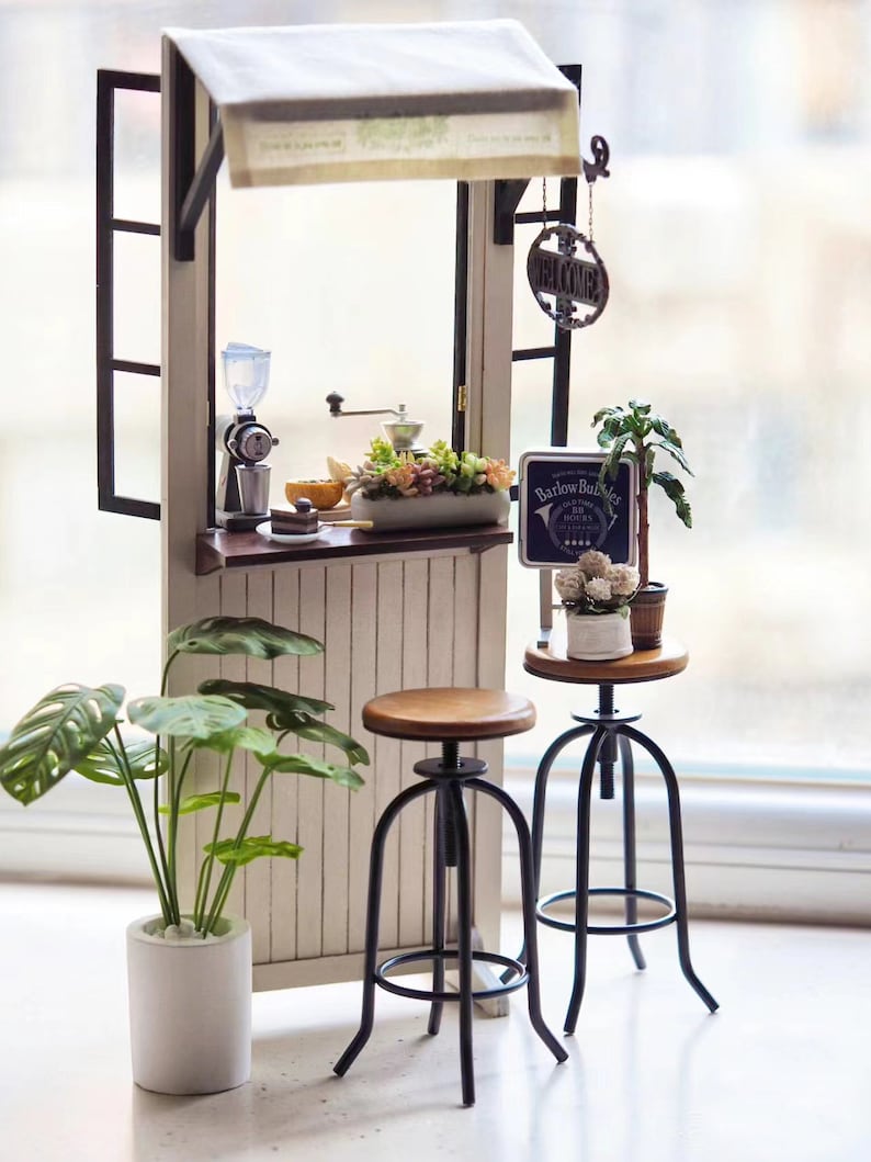 May include: A miniature dollhouse coffee shop with a white awning, two black metal bar stools, and a sign that reads "Barlow Bubbles, 8am - 5pm, The Espresso Bar".