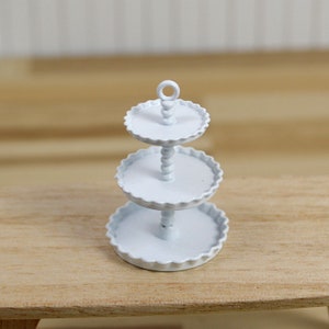 Può includere: Un portavivande bianco a tre livelli con un bordo a festoni su ogni livello. Il portavivande ha una colonna di perline e un manico ad anello nella parte superiore.