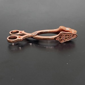May include: A pair of rose gold-colored tongs with an ornate design on the handles. The tongs are closed and lying on a black surface.