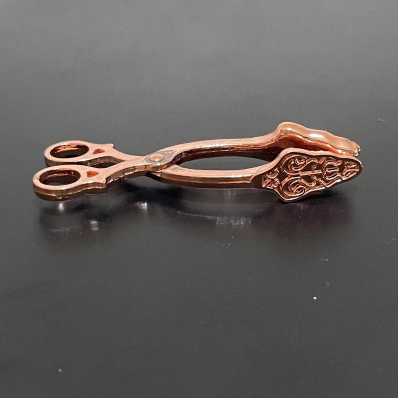 May include: A pair of rose gold-colored tongs with an ornate design on the handles. The tongs are closed and lying on a black surface.
