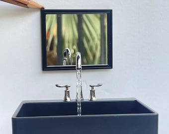 May include: A black rectangular bathroom sink with a chrome faucet and two handles. The faucet is turned on and water is flowing from the spout. A black framed mirror is mounted above the sink.