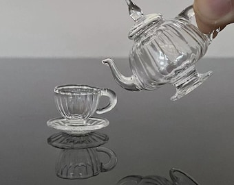May include: A miniature glass tea set with a clear teapot and a cup and saucer. The teapot has a fluted design and a lid with a finial. The cup and saucer are also fluted.