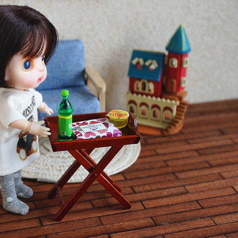 May include: A miniature wooden folding table with a green bottle, a box of chocolates, and a small bowl of yellow jelly on top. The table is set on a brown wooden floor.