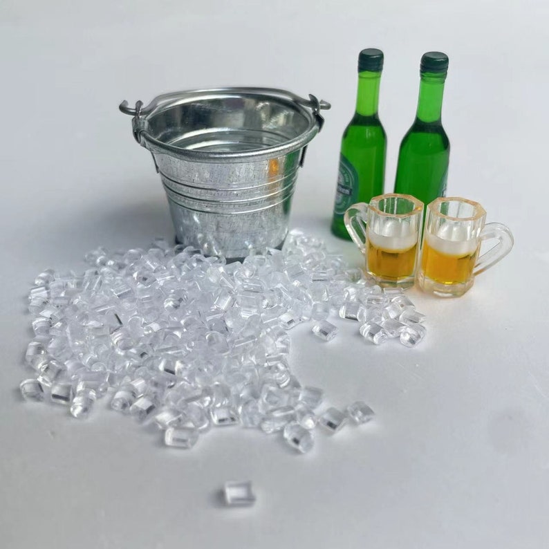 May include: A miniature metal bucket with a handle, two green glass bottles, two clear glass beer mugs filled with a light yellow liquid, and a pile of clear plastic ice cubes.