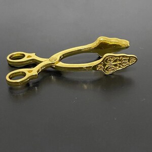 May include: Gold-toned metal tongs with a decorative design on the handles. The tongs are closed and have a slight curve to them.