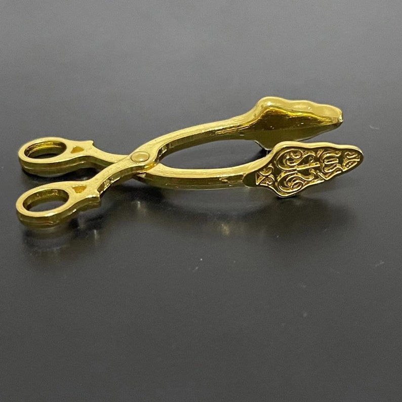 May include: Gold-toned metal tongs with a decorative design on the handles. The tongs are closed and have a slight curve to them.