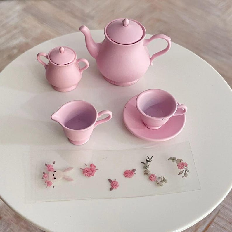 May include: A pink tea set with a teapot, creamer, sugar bowl, and teacup and saucer. The set is made of ceramic and has a delicate floral design.